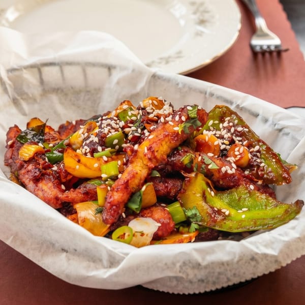 Spicy chicken with peppers and sesame seeds in a basket, with a drink and plate in the background.