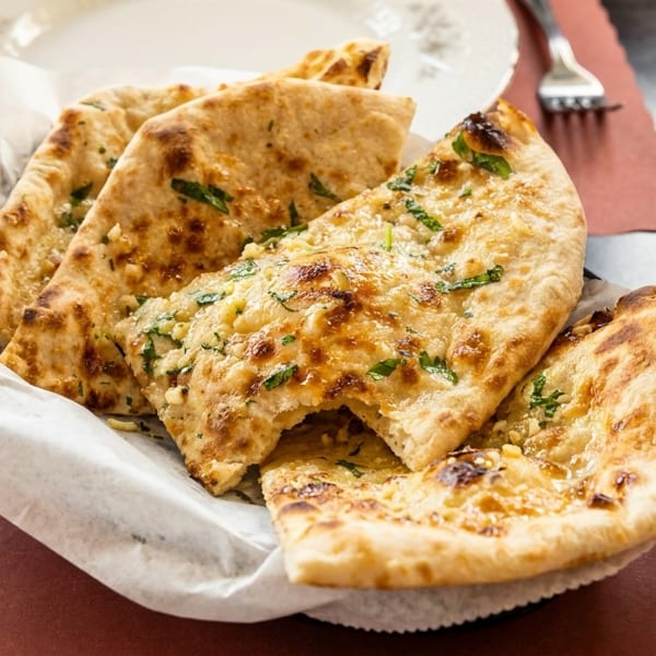 Basket of golden-brown garlic naan bread, garnished with green herbs, on a table with a drink.