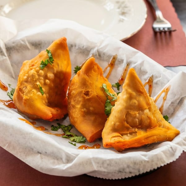 Three golden-brown samosas drizzled with sauce and garnished with cilantro in a white paper-lined basket.