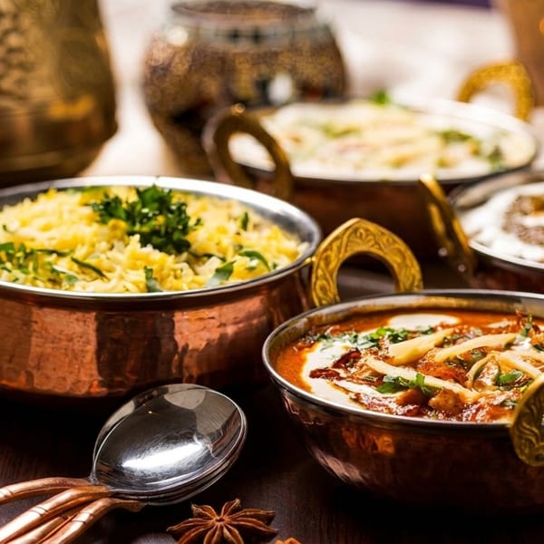Four copper bowls filled with Indian dishes: rice, curries, and raita, on a dark wooden table.