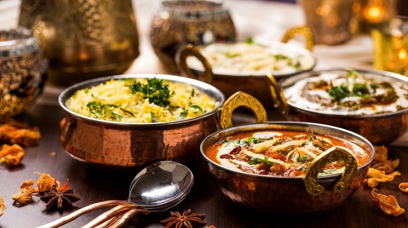 Four copper bowls filled with Indian dishes: rice, curries, and raita, on a dark wooden table.