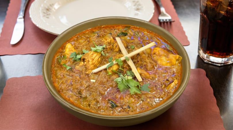 A bowl of chicken curry with cilantro and bamboo shoots, next to a drink and plate.