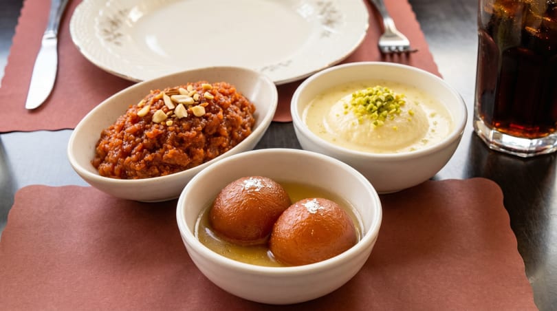 Three bowls of Indian desserts: gulab jamun, rasmalai, and gajar ka halwa, on a table.